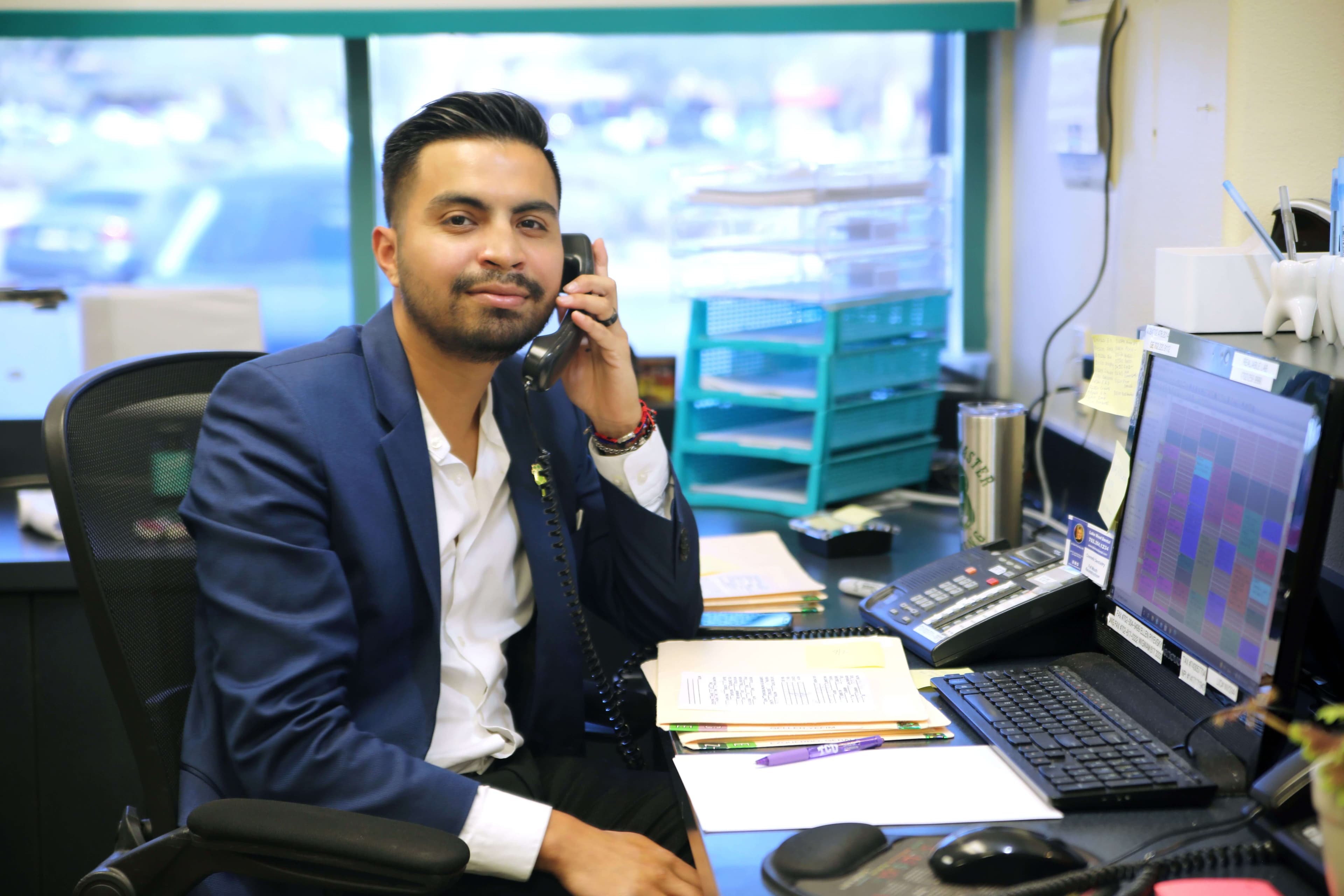 Brunette man sitting in front desc, smiling, holding a phone, welcoming people to contact an office