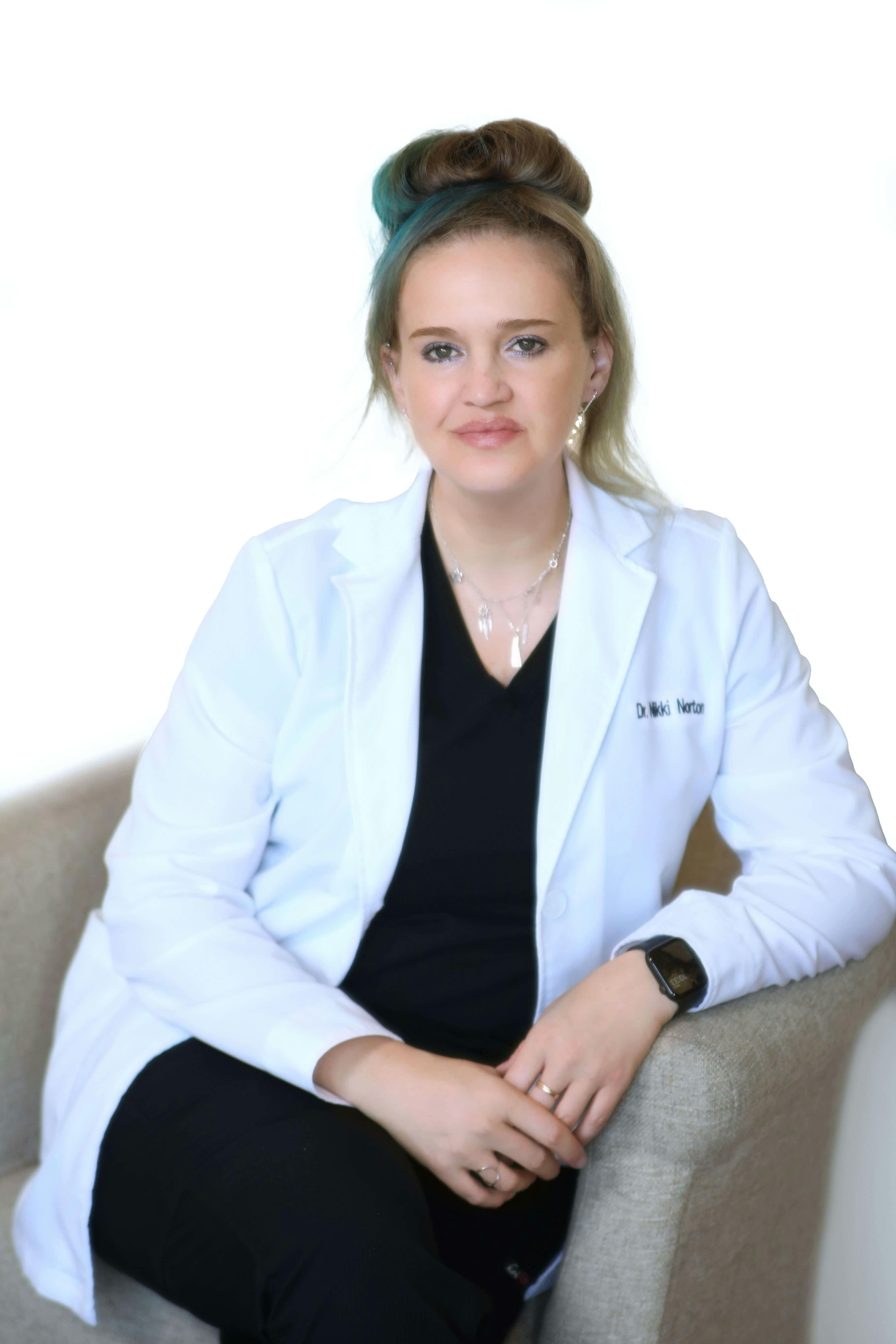 Doctor Nikki Norton, Blonde with blu-ish hair woman, wearing white medical long sleve dress, sitting and smiling